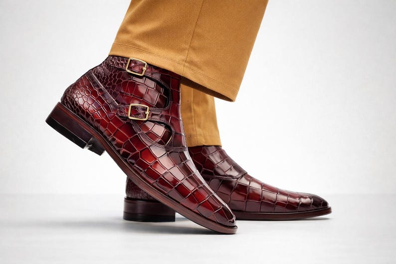 May include: Burgundy leather ankle boots with a crocodile texture and double buckle fastening. The boots have a polished finish and a dark brown sole. The footwear is paired with tan trousers.