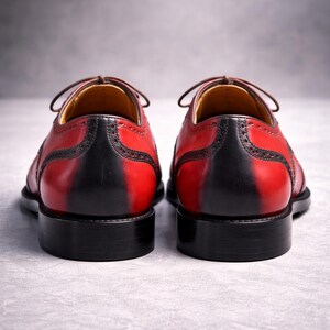 May include: A pair of red and black leather dress shoes. The shoes feature a two-tone design, with red uppers and black heels and soles. Brown laces and stitching are visible. The shoes are set against a grey background.
