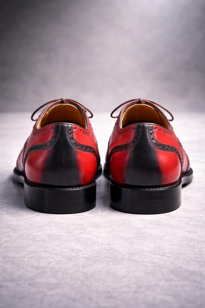 May include: A pair of red and black leather dress shoes. The shoes feature a two-tone design, with red uppers and black heels and soles. Brown laces and stitching are visible. The shoes are set against a grey background.