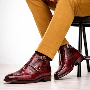 May include: Burgundy leather ankle boots with a crocodile texture. The boots have double straps with gold buckles and a side zip. The boots are worn with mustard-coloured trousers.