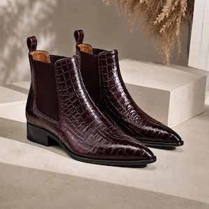 Peut inclure: Une paire de bottines Chelsea bordeaux foncé avec une texture en cuir gaufré façon crocodile, des bouts pointus, des petits talons blocs et des empiècements latéraux élastiques marron foncé avec des tirettes à l'arrière pour un enfilage facile.