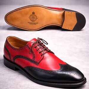 May include: Red and black leather dress shoes with brown laces. The shoes feature a wingtip design and decorative perforations. The sole is visible, showing the brand's logo and the words "Handmade in England".