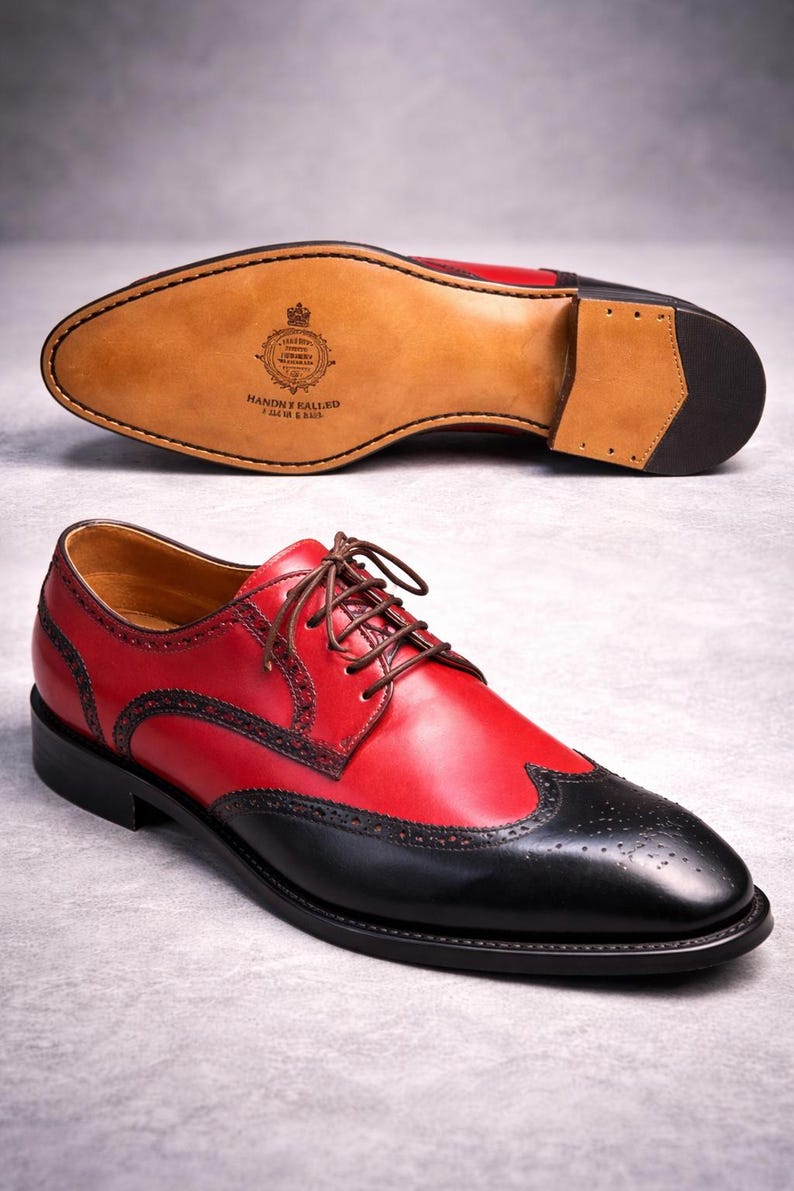 May include: Red and black leather dress shoes with brown laces. The shoes feature a wingtip design and decorative perforations. The sole is visible, showing the brand's logo and the words "Handmade in England".