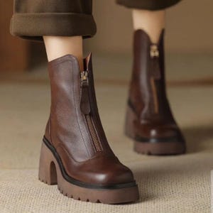 May include: Brown leather ankle boots with a front zip and a chunky, stacked heel. The boots have a rounded toe and a lug sole. The heel is approximately 7.6 cm high. The boots are a rich, warm brown colour.