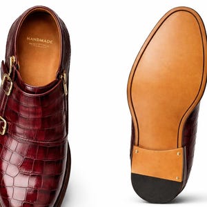 May include: Burgundy leather double monk strap shoes with gold buckles. The shoes feature a textured, crocodile-like pattern and a leather sole. The interior is tan with the word "HANDMADE" printed on the insole.