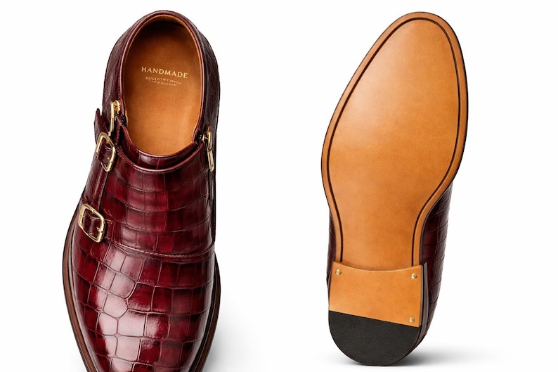 May include: Burgundy leather double monk strap shoes with gold buckles. The shoes feature a textured, crocodile-like pattern and a leather sole. The interior is tan with the word "HANDMADE" printed on the insole.
