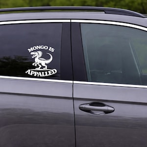 May include: White vinyl car decal featuring a dinosaur illustration with the text "MONGO IS APPALLED". The design is applied to the window of a gray car. The decal is a humorous accessory.