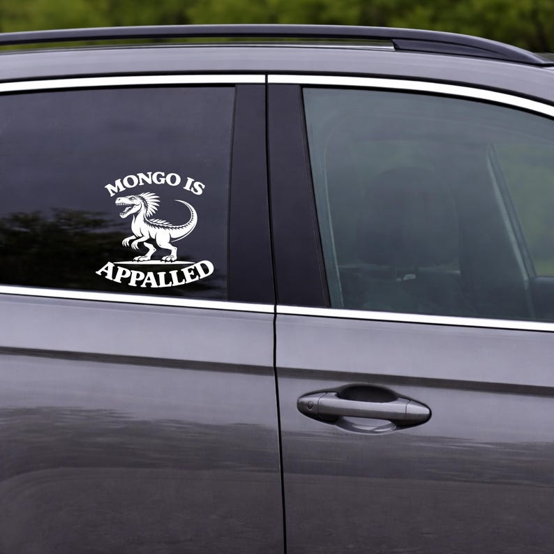 May include: White vinyl car decal featuring a dinosaur illustration with the text "MONGO IS APPALLED". The design is applied to the window of a gray car. The decal is a humorous accessory.