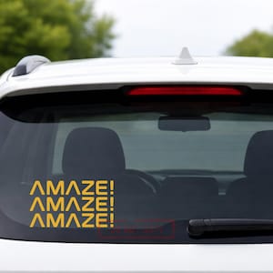 May include: A yellow vinyl car decal with the word "AMAZE!" repeated three times. The decal is applied to the rear window of a white car. The text is in a bold, sans-serif font.