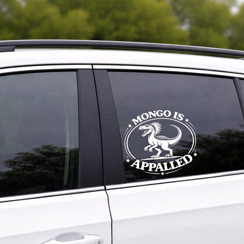 May include: White vinyl decal applied to a car window. The decal features a dinosaur illustration inside an oval with the words "MONGO IS APPALLED" in a stylized font. The background is blurred green.