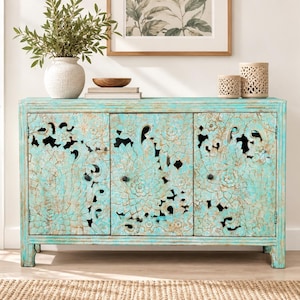 Puede incluir: Un aparador rectangular de madera con tres puertas que presentan intrincados patrones florales cortados con láser. El mueble tiene un acabado de pintura turquesa desgastada y envejecida que revela tonos de madera natural, y tres pequeños pomos circulares oscuros.