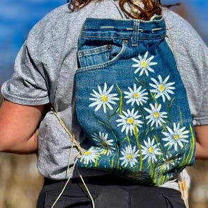 Peut inclure: Un sac à dos en jean avec un motif floral peint de marguerites blanches à cœur jaune et tiges vertes. Le sac à dos est fait à partir de jeans recyclés et possède une fermeture à cordon et des bretelles réglables.