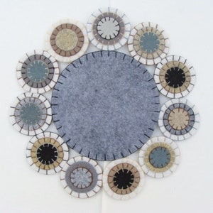 May include: A felt placemat with a large gray center circle, surrounded by twelve smaller, circular coasters. The coasters have a layered design with various neutral colors, including white, gray, and beige, and are stitched with black thread.