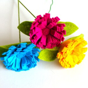 May include: Three felt flowers in vibrant colors: fuchsia, blue, and yellow. Each flower has a layered, textured design, with green felt leaves. The flowers are attached to a green stem, creating a cheerful, handmade floral arrangement.