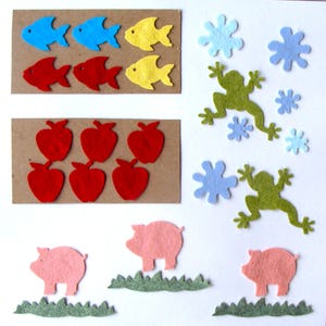 May include: A collection of felt cutouts featuring various shapes and colors. The image includes blue, red, and yellow fish, red apples, green frogs, blue snowflakes, and pink pigs with green grass.