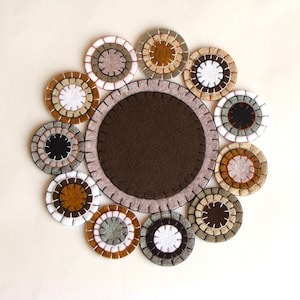 May include: A felt coaster set featuring a large brown center mat surrounded by smaller, circular coasters. The coasters have concentric rings of various colors, including brown, white, and gray, with stitched edges. The set is arranged on a cream-colored surface.