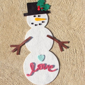 May include: A white felt snowman decoration with a black top hat, orange carrot nose, and brown stick arms. The snowman has a green holly and red berry accent on the hat, a blue heart, and the word "love" in pink.