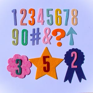 May include: A collection of colorful felt numbers, symbols, and shapes. The numbers 1 through 9, 0, and symbols like the hashtag, ampersand, and question mark are displayed. There is also an upward arrow, a flower with the number 3, a star with the number 5, and a ribbon with the number 2.