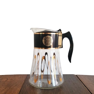 Puede incluir: Una cafetera de vidrio negro y dorado con un asa de plástico negro. La cafetera tiene un diseño dorado en la parte inferior y un medallón dorado en la tapa.