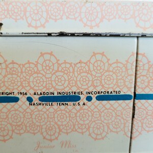 Peut inclure: Une bo&icirc;te &agrave; lunch en m&eacute;tal blanc avec un motif de dentelle rose et le texte "COPYRIGHT 1956 ALADDIN INDUSTRIES, INCORPORATED NASHVILLE, TENN., U.S.A. Junior Miss".