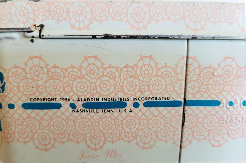 Peut inclure: Une bo&icirc;te &agrave; lunch en m&eacute;tal blanc avec un motif de dentelle rose et le texte "COPYRIGHT 1956 ALADDIN INDUSTRIES, INCORPORATED NASHVILLE, TENN., U.S.A. Junior Miss".