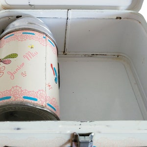 Peut inclure: Bo&icirc;te &agrave; lunch en m&eacute;tal blanc avec une gourde assortie. La gourde est d&eacute;cor&eacute;e d'un motif floral rose et bleu avec les mots "Junior Miss" dessus.