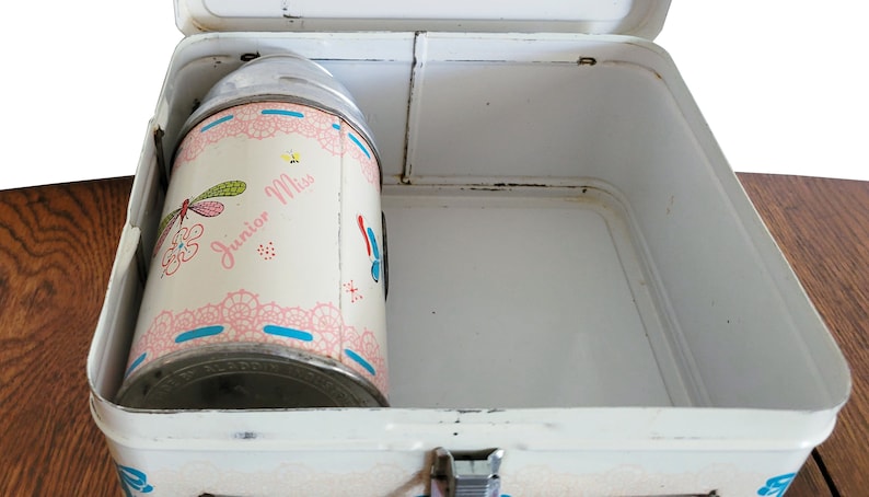 Peut inclure: Bo&icirc;te &agrave; lunch en m&eacute;tal blanc avec une gourde assortie. La gourde est d&eacute;cor&eacute;e d'un motif floral rose et bleu avec les mots "Junior Miss" dessus.