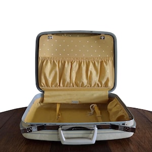 May include: An open vintage suitcase with a light-colored exterior and a bright yellow interior lining. The interior features a polka-dot pattern on the lid panel, an elasticized storage pocket, and a white plastic handle on the front.