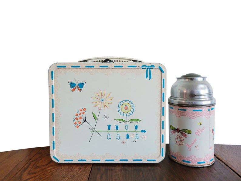 Peut inclure: Ensemble vintage de bo&icirc;te &agrave; lunch et de thermos en m&eacute;tal avec un motif floral. La bo&icirc;te &agrave; lunch pr&eacute;sente un motif floral bleu et blanc avec un papillon. Le thermos a un motif floral rose et bleu avec une libellule et le texte "Miss Junior".