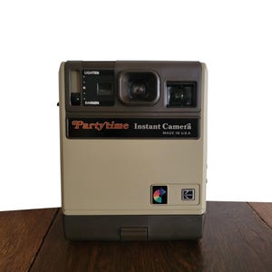 May include: A beige and brown Partytime Instant Camera with a black lens and a light/darken dial. The camera is made in the USA.