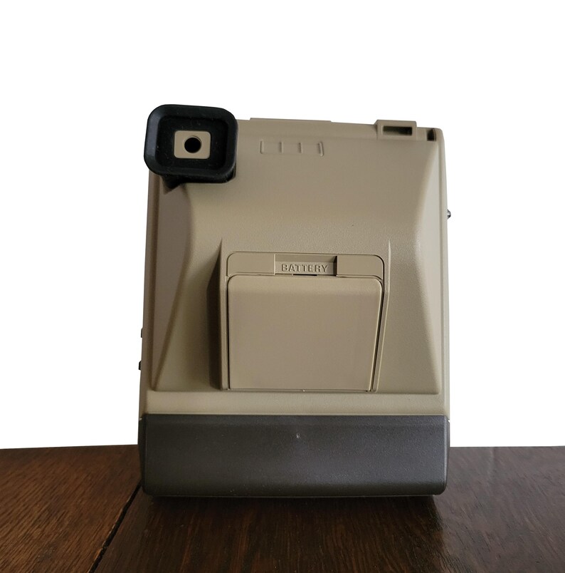 May include: A beige instant camera with a black lens and a battery compartment labeled "BATTERY".