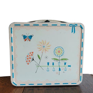 Peut inclure: Bo&icirc;te &agrave; lunch vintage en m&eacute;tal avec un fond blanc et un motif floral bleu, rose et jaune. La bo&icirc;te &agrave; lunch a un ruban bleu et une poign&eacute;e blanche.