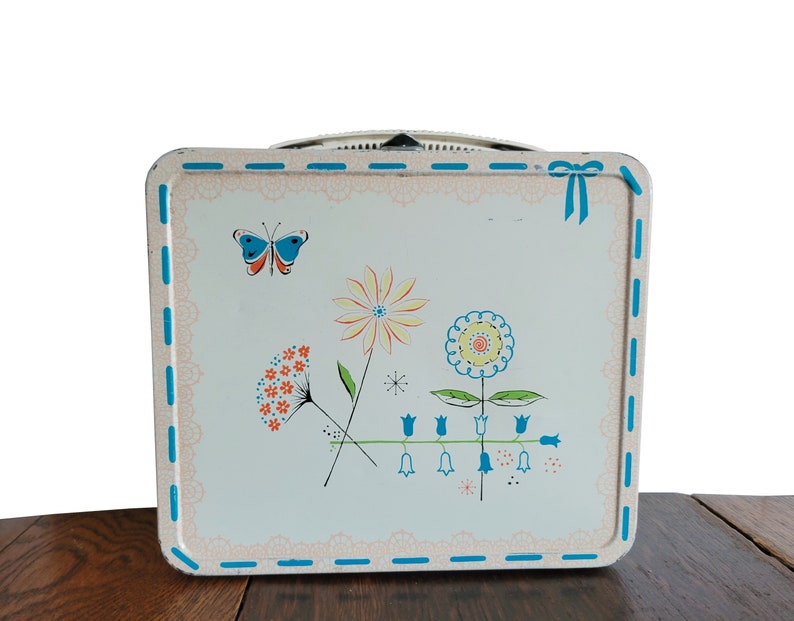 Peut inclure: Bo&icirc;te &agrave; lunch vintage en m&eacute;tal avec un fond blanc et un motif floral bleu, rose et jaune. La bo&icirc;te &agrave; lunch a un ruban bleu et une poign&eacute;e blanche.