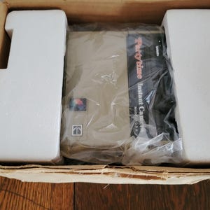 May include: A white cardboard box with a brown interior. The box contains a Party Time Instant Camera, wrapped in plastic. The camera is beige with a black front panel that says "Party Time Instant Camera Made in USA".