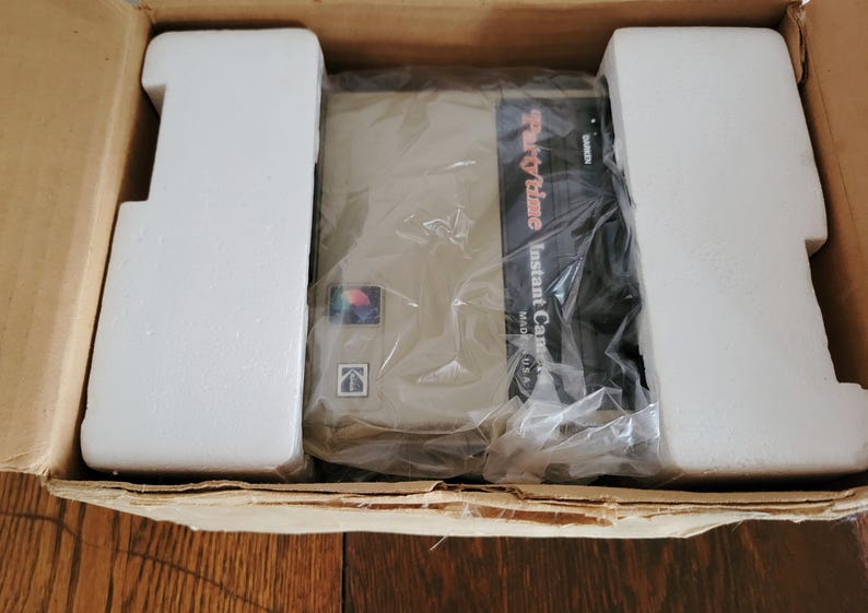 May include: A white cardboard box with a brown interior. The box contains a Party Time Instant Camera, wrapped in plastic. The camera is beige with a black front panel that says "Party Time Instant Camera Made in USA".