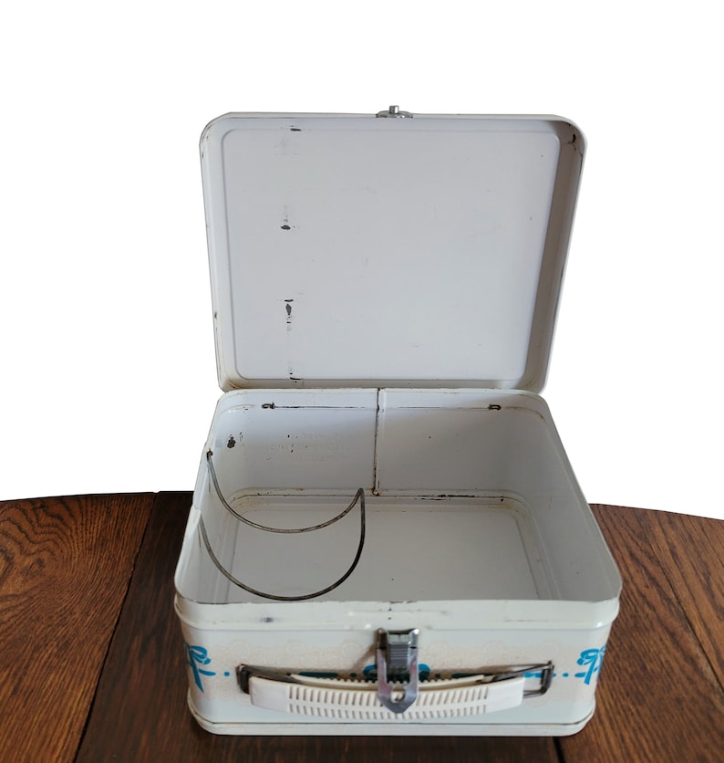 Peut inclure: Bo&icirc;te &agrave; lunch en m&eacute;tal blanc avec un motif bleu et blanc. La bo&icirc;te &agrave; lunch est ouverte et l'int&eacute;rieur est vide. Il y a une poign&eacute;e en m&eacute;tal sur le c&ocirc;t&eacute; de la bo&icirc;te &agrave; lunch.