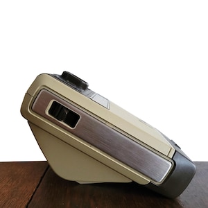 May include: A beige and brown vintage camera with a silver accent on the side. The camera is sitting on a wooden surface.