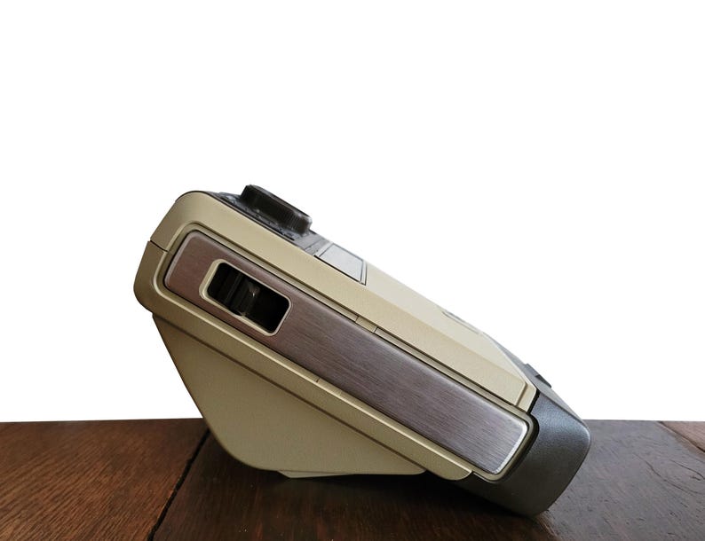 May include: A beige and brown vintage camera with a silver accent on the side. The camera is sitting on a wooden surface.