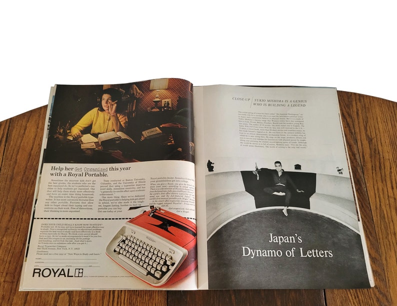 Op de afbeelding: Een dubbele pagina uit een vintage tijdschrift met een advertentie voor een Royal Portable typemachine. De advertentie toont een vrouw die aan een bureau werkt met boeken en een lamp. De typemachine is rood en wit en heeft de tekst "Help haar dit jaar georganiseerd te zijn met een Royal Portable." De andere pagina toont een zwart-witfoto van een man die op een gebogen platform zit met de tekst "Close-up: Fukio Mishima, een genie dat een legende bouwt." De tekst "Japans Dynamo van Letters" is ook te zien.