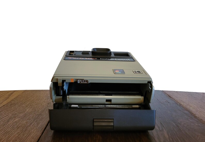May include: A vintage Polaroid camera with a gray and black exterior. The camera is open, revealing the film compartment and a pull tab labeled "PULL".