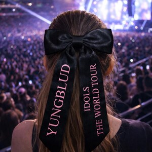 May include: A black satin hair bow with pink text reading "YUNGBLUD" and "IDOLS THE WORLD TOUR". The bow is worn in the hair of a person at a concert, with a crowd and stage lights in the background.
