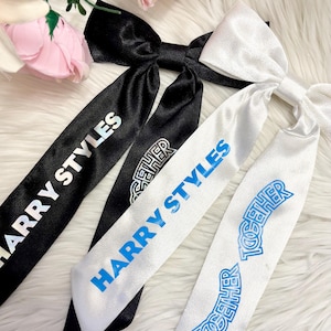 May include: Satin hair bows in black and white, featuring the text "HARRY STYLES" and "TOGETHER" printed in different colours. The bows are displayed on a fluffy white surface, with pink flowers in the background.