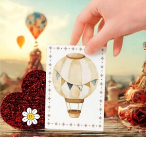 Può includere: Una mano tiene in mano una carta con un'illustrazione ad acquerello di una mongolfiera. Il pallone è beige e bianco con accenti blu e oro. Un cuore rosso glitterato con una margherita è in primo piano, con mongolfiere sfocate sullo sfondo.