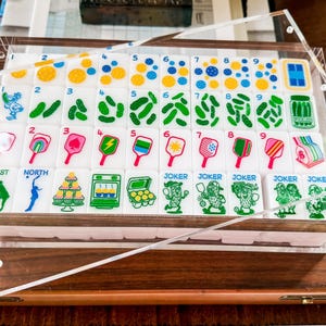 Puede incluir: Un juego de fichas de mahjong rectangulares blancas dispuestas en una bandeja de acr&iacute;lico transparente. Las fichas presentan ilustraciones coloridas que incluyen n&uacute;meros, frascos de pepinillos, paletas de pickleball y personajes joker.