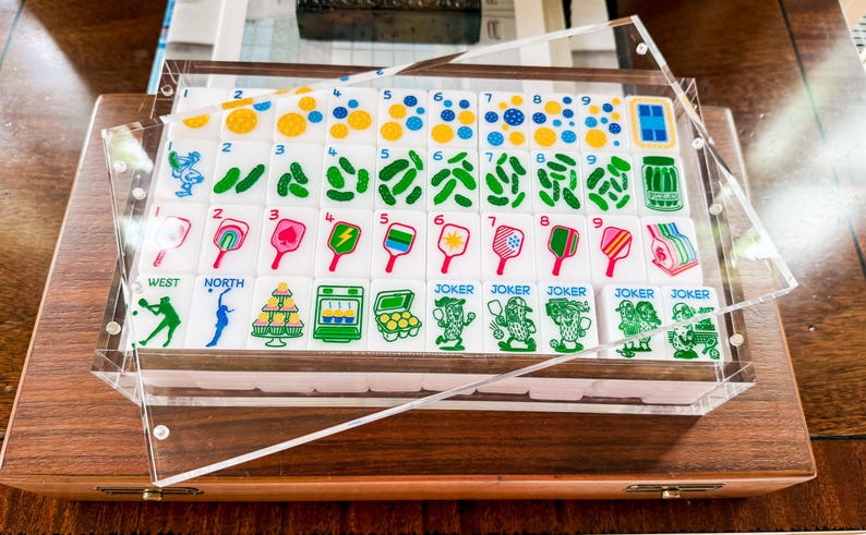 Puede incluir: Un juego de fichas de mahjong rectangulares blancas dispuestas en una bandeja de acr&iacute;lico transparente. Las fichas presentan ilustraciones coloridas que incluyen n&uacute;meros, frascos de pepinillos, paletas de pickleball y personajes joker.