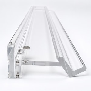 May include: A clear, transparent acrylic wall-mounted display shelf featuring a long, rectangular back panel and a protruding ledge with a raised front lip. The structure is held together by small, circular silver-toned metal hardware fasteners.