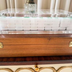 Puede incluir: Una caja de acr&iacute;lico transparente que contiene un juego de fichas de Mahjong blancas con marcas verdes y azules, apoyada sobre un estuche de almacenamiento rectangular de madera con bisagras de metal color lat&oacute;n y acabado en madera marr&oacute;n.