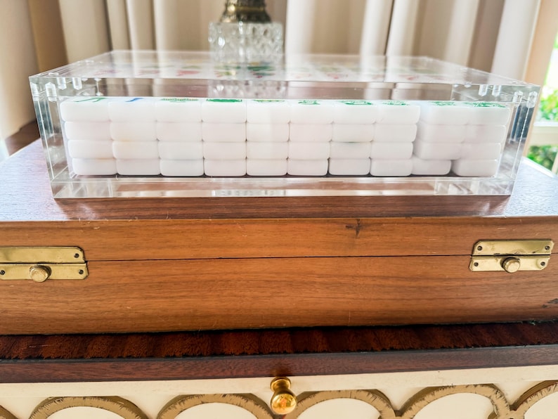 Puede incluir: Una caja de acr&iacute;lico transparente que contiene un juego de fichas de Mahjong blancas con marcas verdes y azules, apoyada sobre un estuche de almacenamiento rectangular de madera con bisagras de metal color lat&oacute;n y acabado en madera marr&oacute;n.