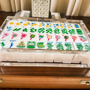 Puede incluir: Un juego de fichas de mahjong blancas con gr&aacute;ficos coloridos de tem&aacute;tica pickleball, que incluyen palas, pelotas y frascos de pepinillos, dentro de una caja de acr&iacute;lico transparente. Las fichas muestran n&uacute;meros del uno al nueve y jokers.