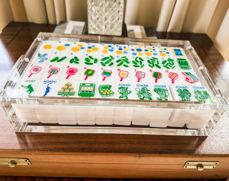 Puede incluir: Un juego de fichas de mahjong blancas con gr&aacute;ficos coloridos de tem&aacute;tica pickleball, que incluyen palas, pelotas y frascos de pepinillos, dentro de una caja de acr&iacute;lico transparente. Las fichas muestran n&uacute;meros del uno al nueve y jokers.